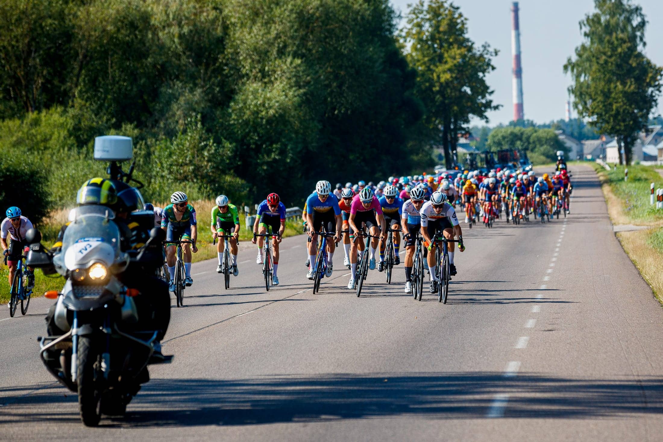 Fahrerfeld auf gerader Landstraße, Begleitmotorrad im Vordergrund, die Straße zieht sich bis zum Horizont